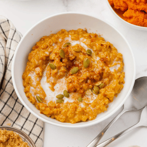 A white bowl filled with pumpkin oatmeal, topped with pepitas. Around the bowl is other bowls of the ingredients used to make the oatmeal.