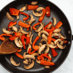 top view photo of a frying pan with mushrooms and red bell peppers, being stirred by a wooden spoon, and cooked until soft and lightly browned