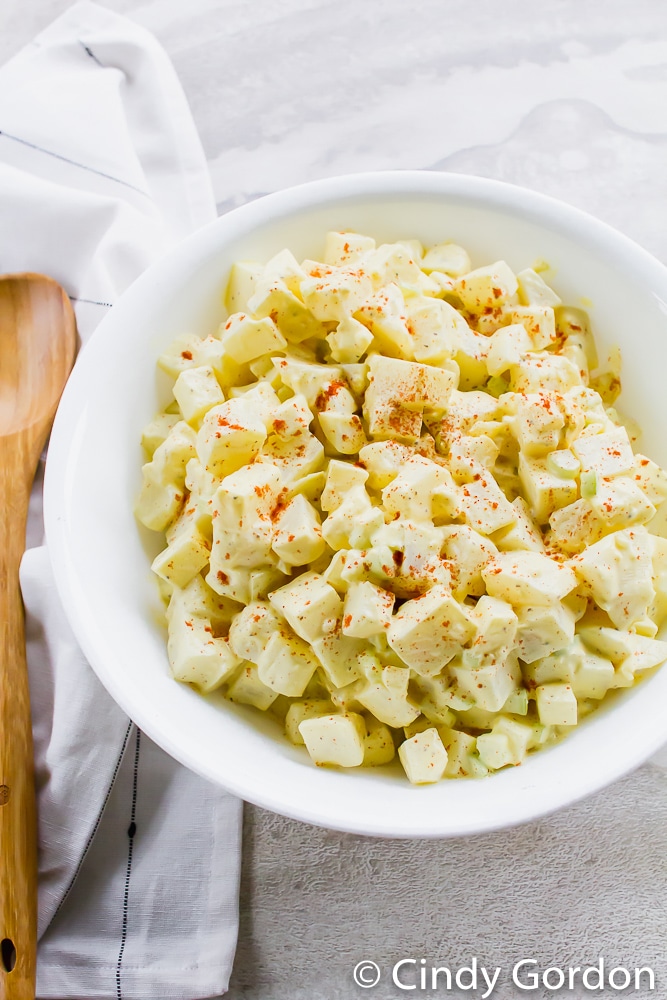 a bowl of amish potato salad with eggs and celery on a counter next to a wooden spoon. 