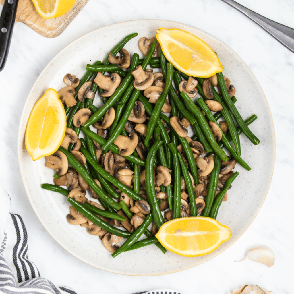 a round white plate filled with green beans and mushrooms. Three lemon wedges are around the edge of the plate.