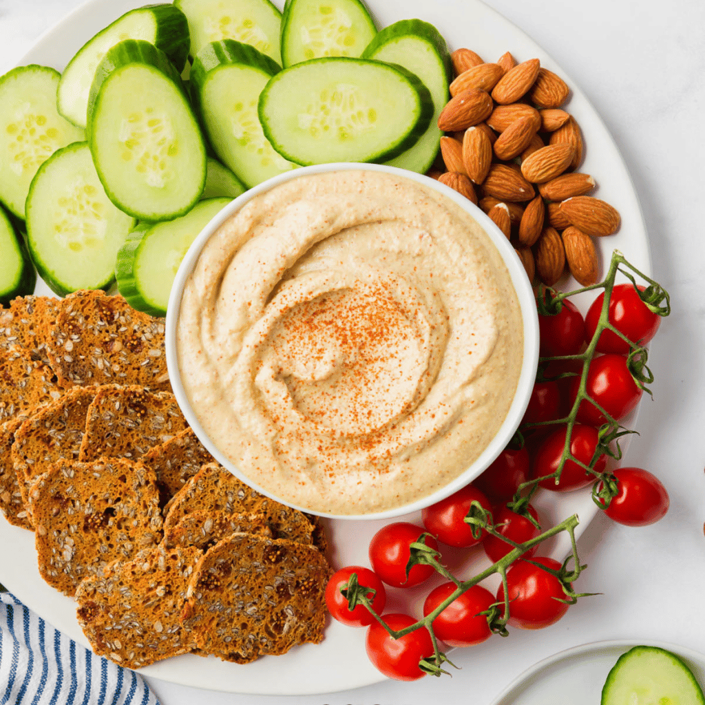 a white bowl of homemade copycat bitchin sauce, swirled with paprika sprinkled on top. almonds, tomatoes, cucumbers, and seed crackers are around the bowl, and you're looking at it from overhead.