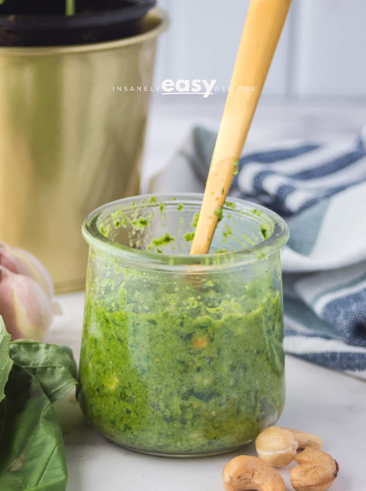 closeup photo of cashew pesto, in a small glass jar with a gold spoon dipped inside. There is a gold pot of basil and a kitchen towel in the background, and basil, cashews, and garlic in the foreground