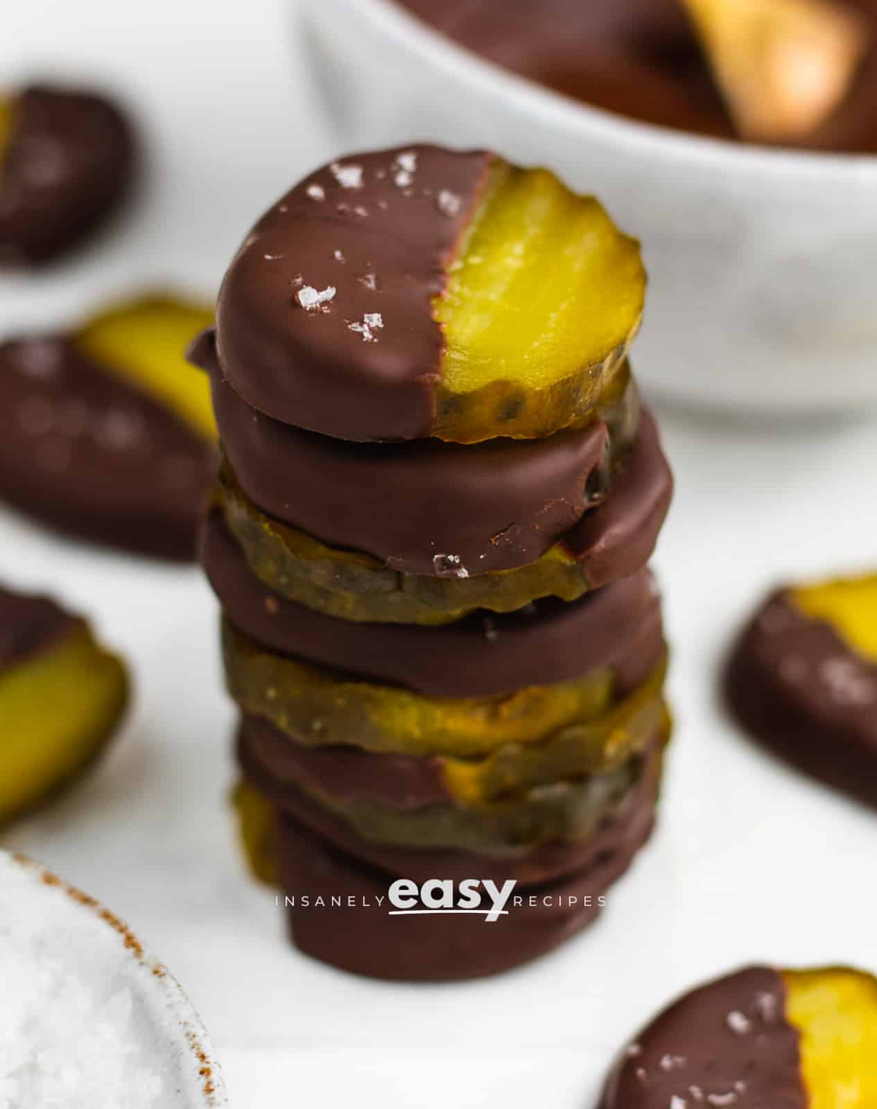 closeup photo of chocolate covered pickles in a stack, with a bowl of flaky salt in the foreground and chocolate covered pickles in the background, and a white bowl of melted chocolate in the background
