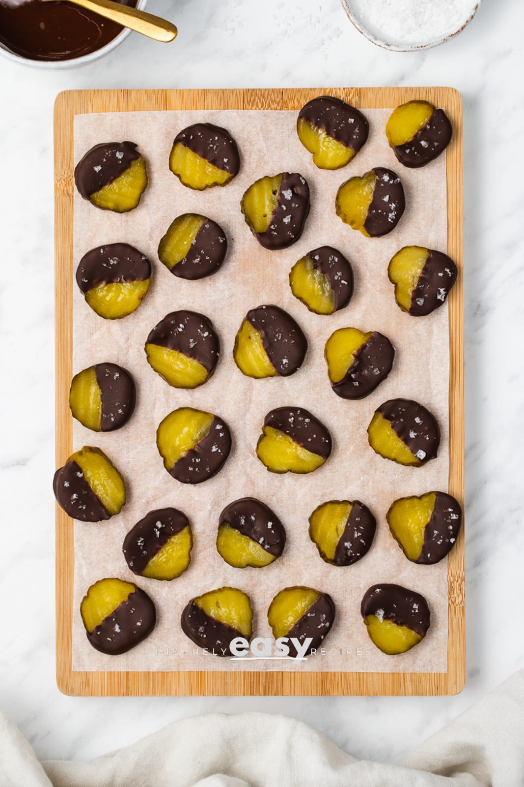 top view photo of pickles dipped in chocolate and sprinkled with salt, on a sheet of parchment paper on a wooden cutting board, with a white bowl of melted chocolate and a white bowl of salt above the board, and a white kitchen towel below the board