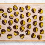 top view photo of pickles dipped in chocolate and sprinkled with salt, on a sheet of parchment paper on a wooden cutting board, with a white bowl of melted chocolate and a white bowl of salt to the right of the board