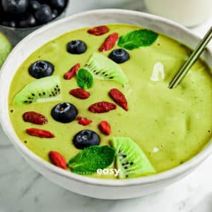 closeup photo of a Matcha Bowl with sliced kiwis, goji berries, blueberries, and mint leaves on top. There is a gold spoon in the bowl and blueberries in the background