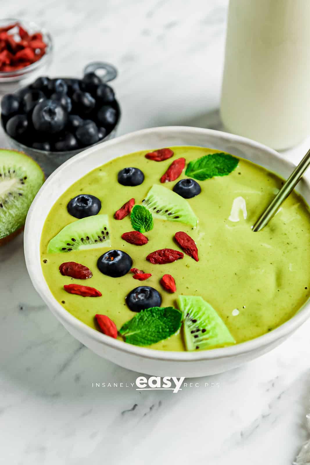 photo of a Matcha Bowl with sliced kiwis, goji berries, blueberries, and mint leaves on top. There is a gold spoon in the bowl and blueberries, half a kiwi, goji berries, and a jar of coconut milk in the background