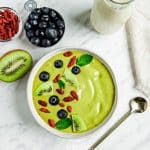 top view photo of a Matcha Bowl with sliced kiwis, goji berries, blueberries, and mint leaves on top. There is a gold spoon below the bowl and blueberries, half a kiwi, goji berries, and a jar of coconut milk above the bowl.
