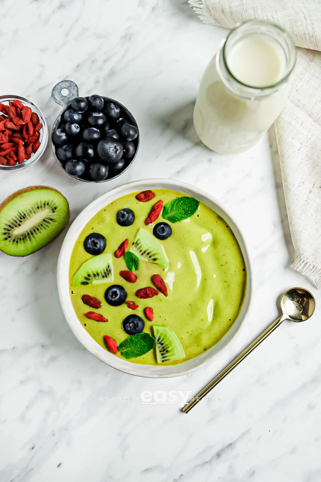 top view photo of a Matcha Bowl with sliced kiwis, goji berries, blueberries, and mint leaves on top. There is a gold spoon below the bowl and blueberries, half a kiwi, goji berries, and a jar of coconut milk above the bowl.