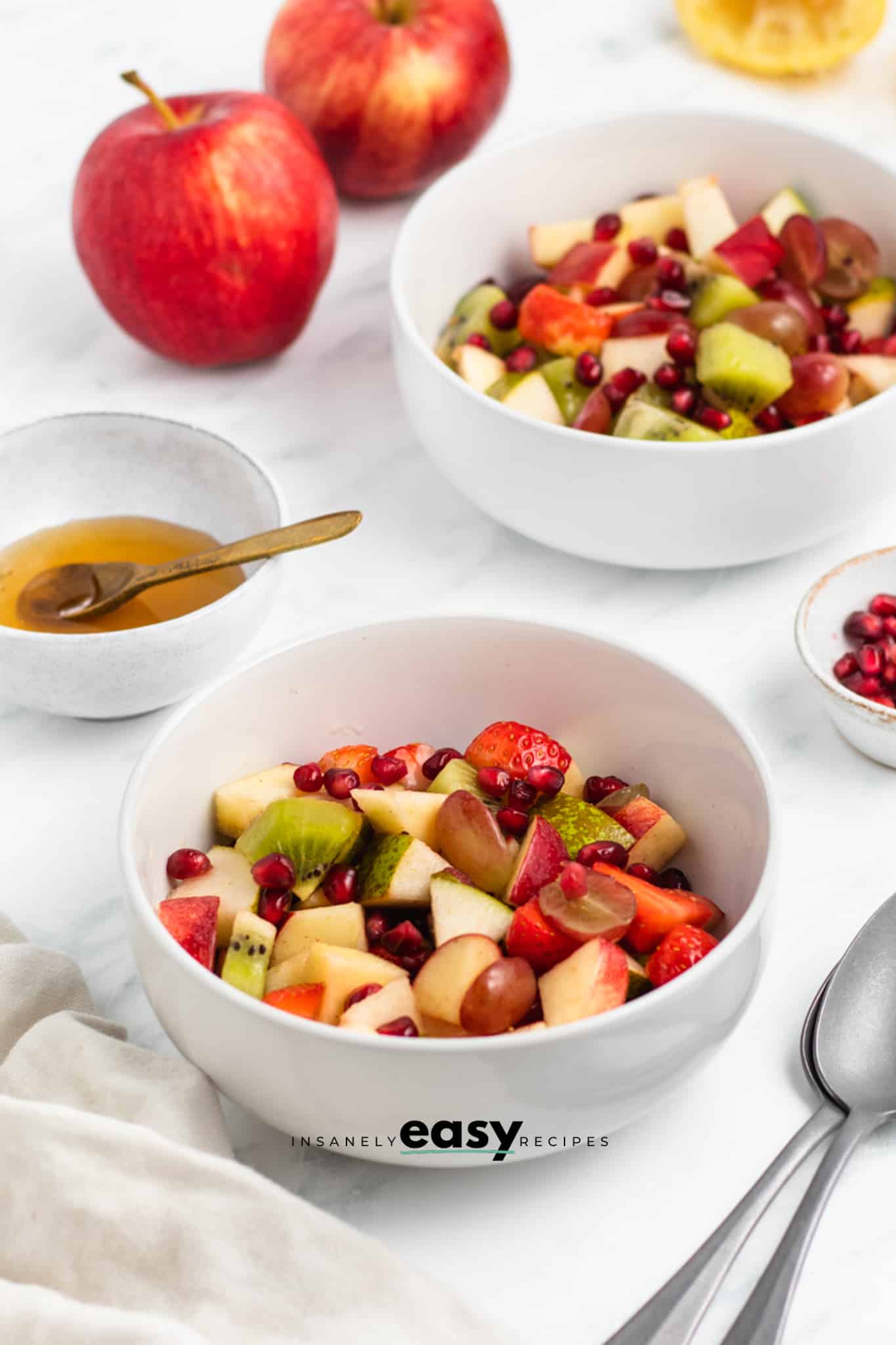 Photo of 2 servings of Thanksgiving Fruit Salad in two white bowls. There is a white bowl with honey and a small white bowl with pomegranate seeds by the salad. And there are two red apples in the background.  