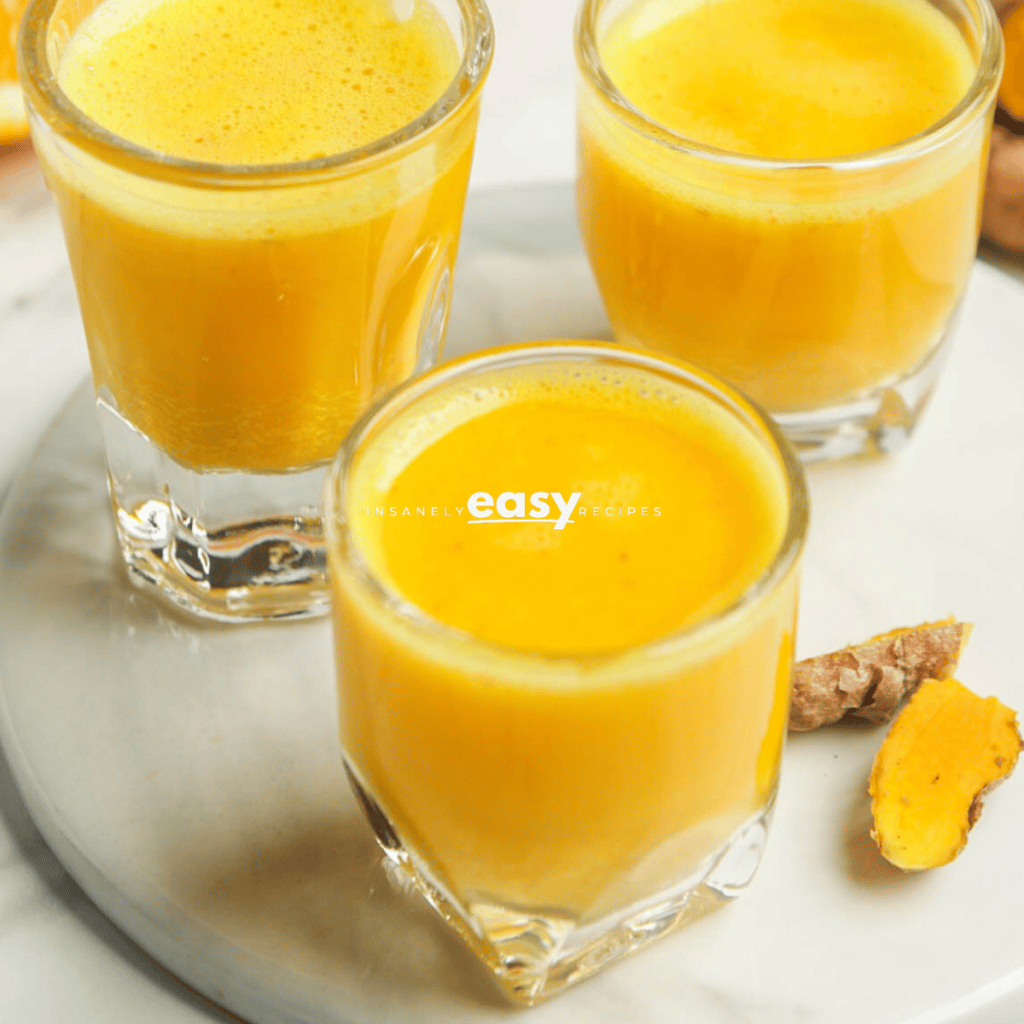 closeup photo of 3 shot glasses filled with turmeric shots. They are on a white plate with fresh turmeric on the plate.