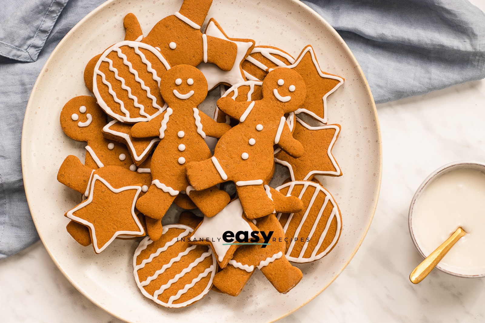 top view photo of a plate of vegan gingerbread cookies with a bowl of vegan icing to the right with a gold spoon. There is a blue kitchen towel above the white plate. 