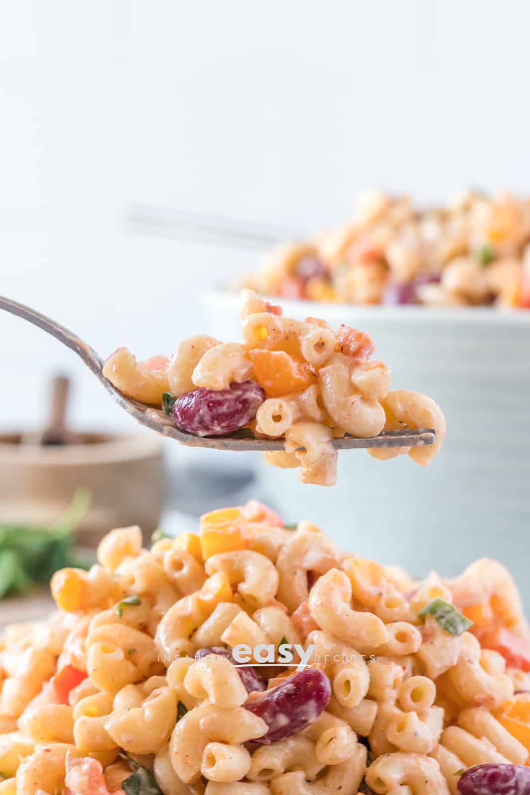 Closeup photo of a fork scooping up Vegan Macaroni Salad from a bowl. There is a white bowl with Vegan Macaroni Salad in the background.