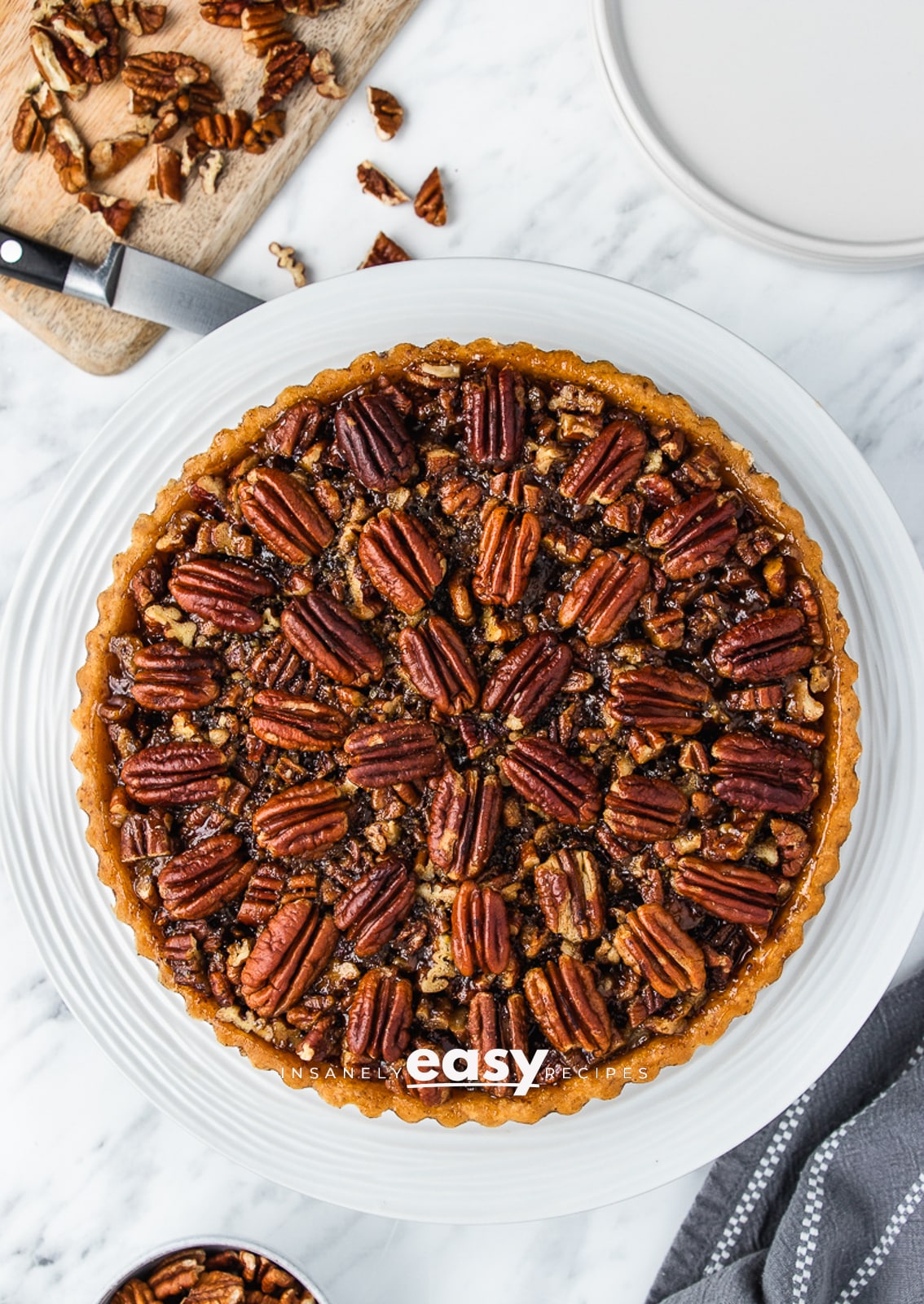 top view photo of vegan pecan pie with chopped pecans and knife above the pie, and a blue kitchen towel to the bottom of the pie
