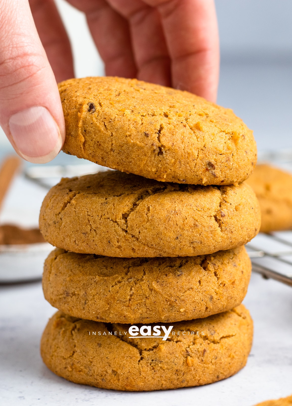 Photo of a stack of 4 Vegan Pumpkin Cookies, with a hand grabbing the cookie on top.