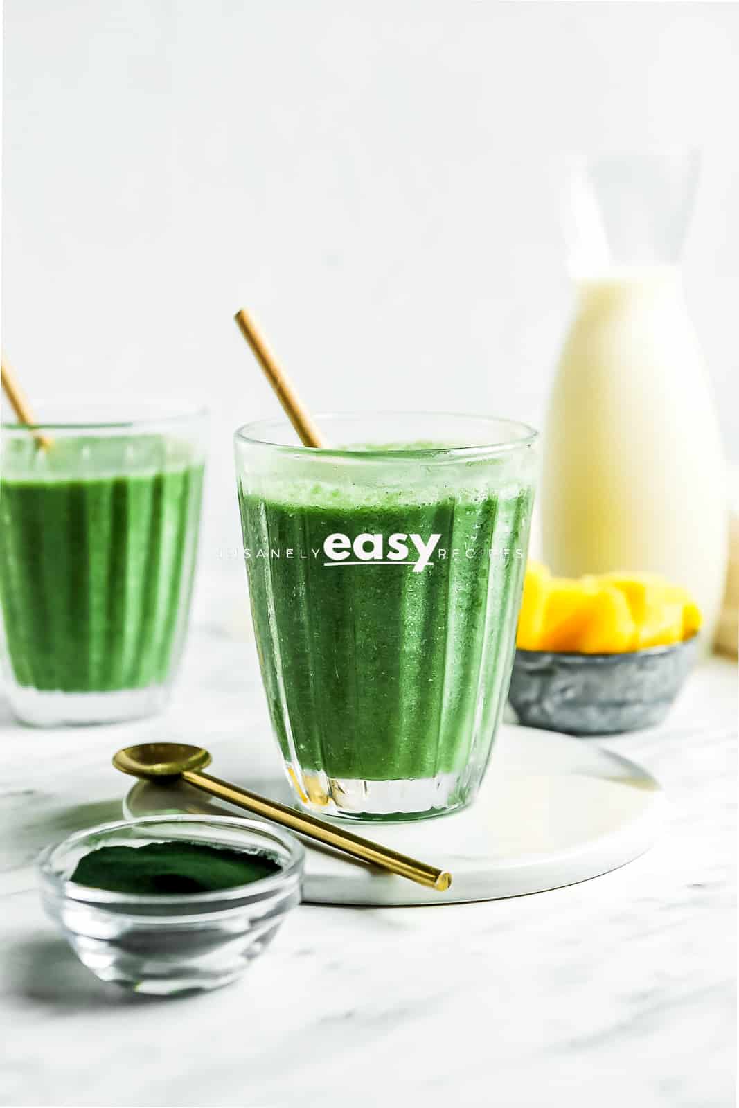 Photo of Spirulina Smoothie in a small glass on a white plate. with a gold spoon. There is another glass with smoothie in the background as well as frozen pineapple, plant milk, and spirulina powder in containers. 
