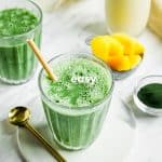 Closeup photo of Spirulina Smoothie in a glass jar with a paper straw, on a white plate with a gold spoon. There are frozen pineapples in the background and another glass of smoothie.