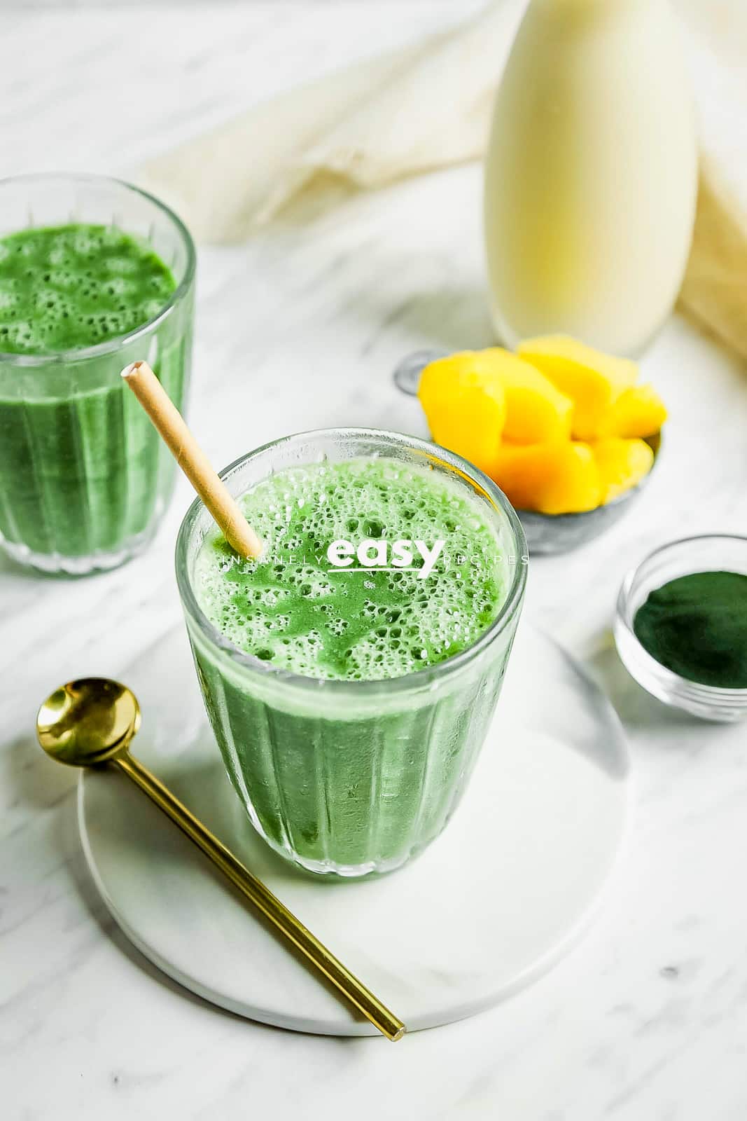 Closeup photo of Spirulina Smoothie in a glass jar with a paper straw, on a white plate with a gold spoon. There are frozen pineapples in the background and another glass of smoothie.