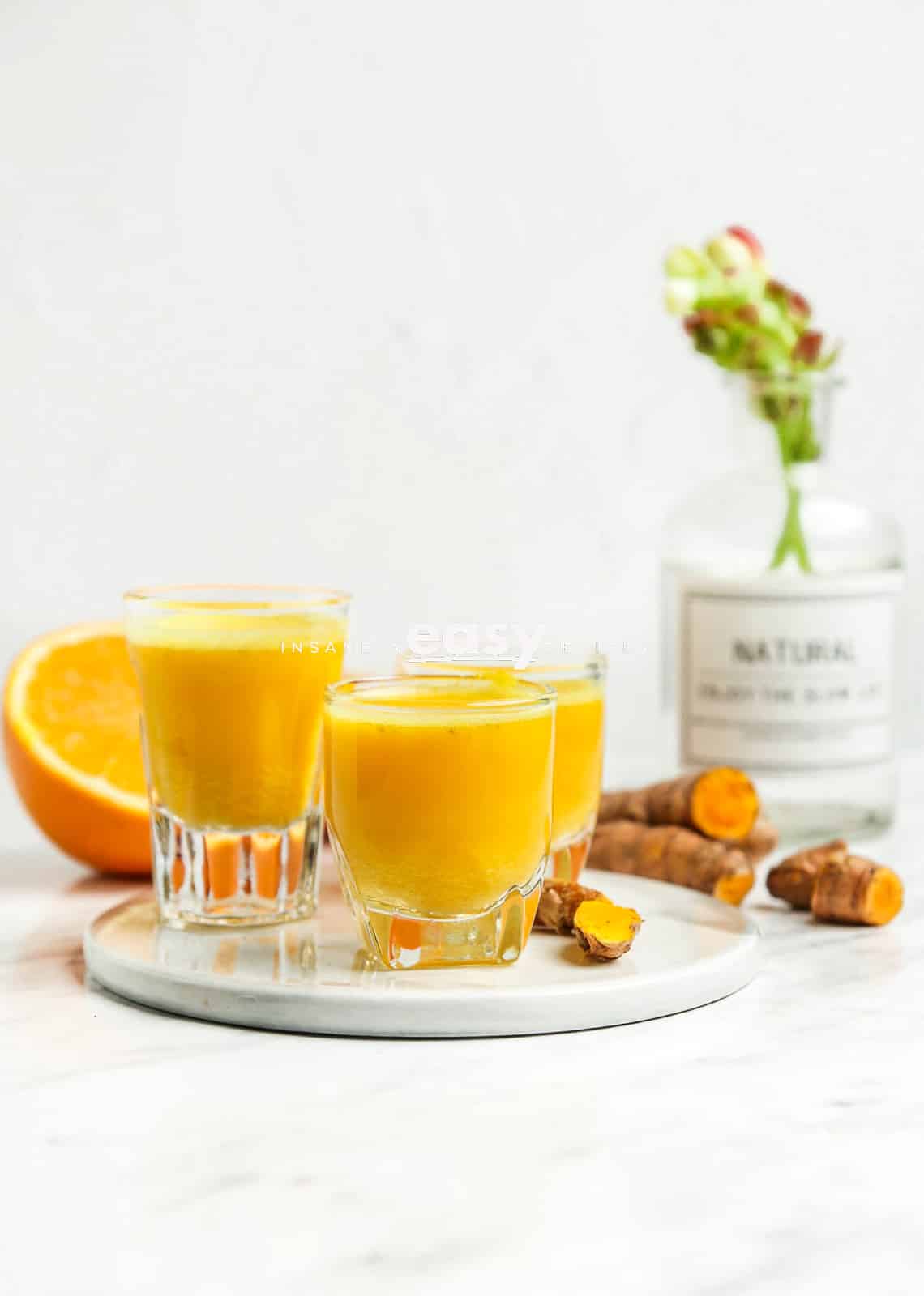 Photo of 3 shot glasses filled with turmeric shots, on a white plate.  There are sliced oranges, fresh turmeric, and flowers in a jar in the background. 