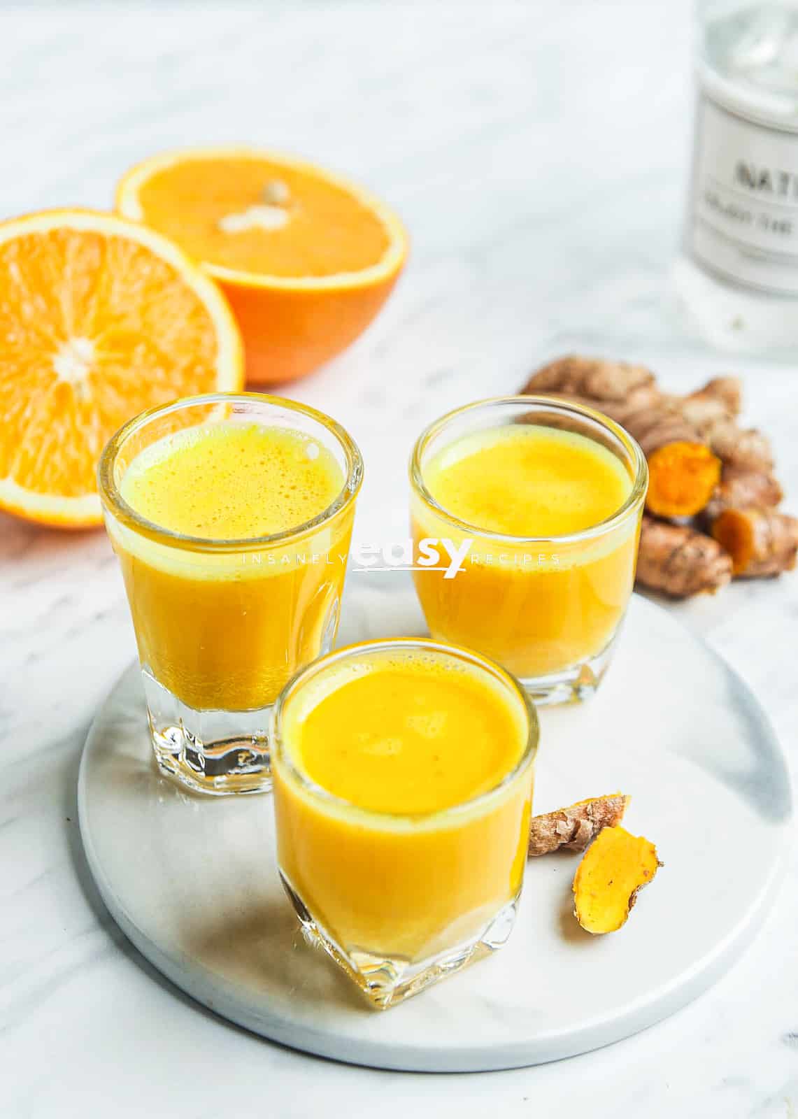 photo of 3 shot glasses filled with turmeric shots, on a white plate, with slices of fresh oranges and fresh turmeric in the background.
