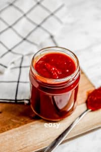Photo of Vegan BBQ Sauce in a clear jar with a silver spoon with sauce next to the jar. It is sitting on a wooden cutting board with a kitchen towel in the background.