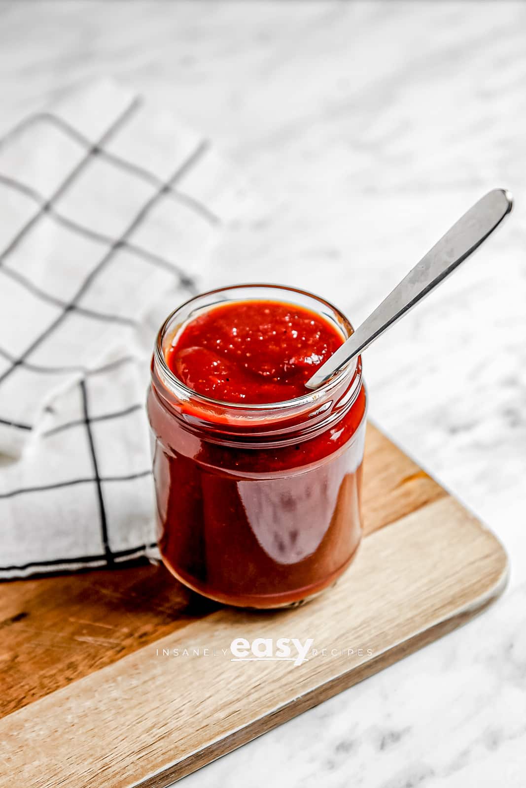 Photo of Vegan BBQ Sauce in a clear jar with a silver spoon in the jar. It is sitting on a wooden cutting board with a kitchen towel in the background.