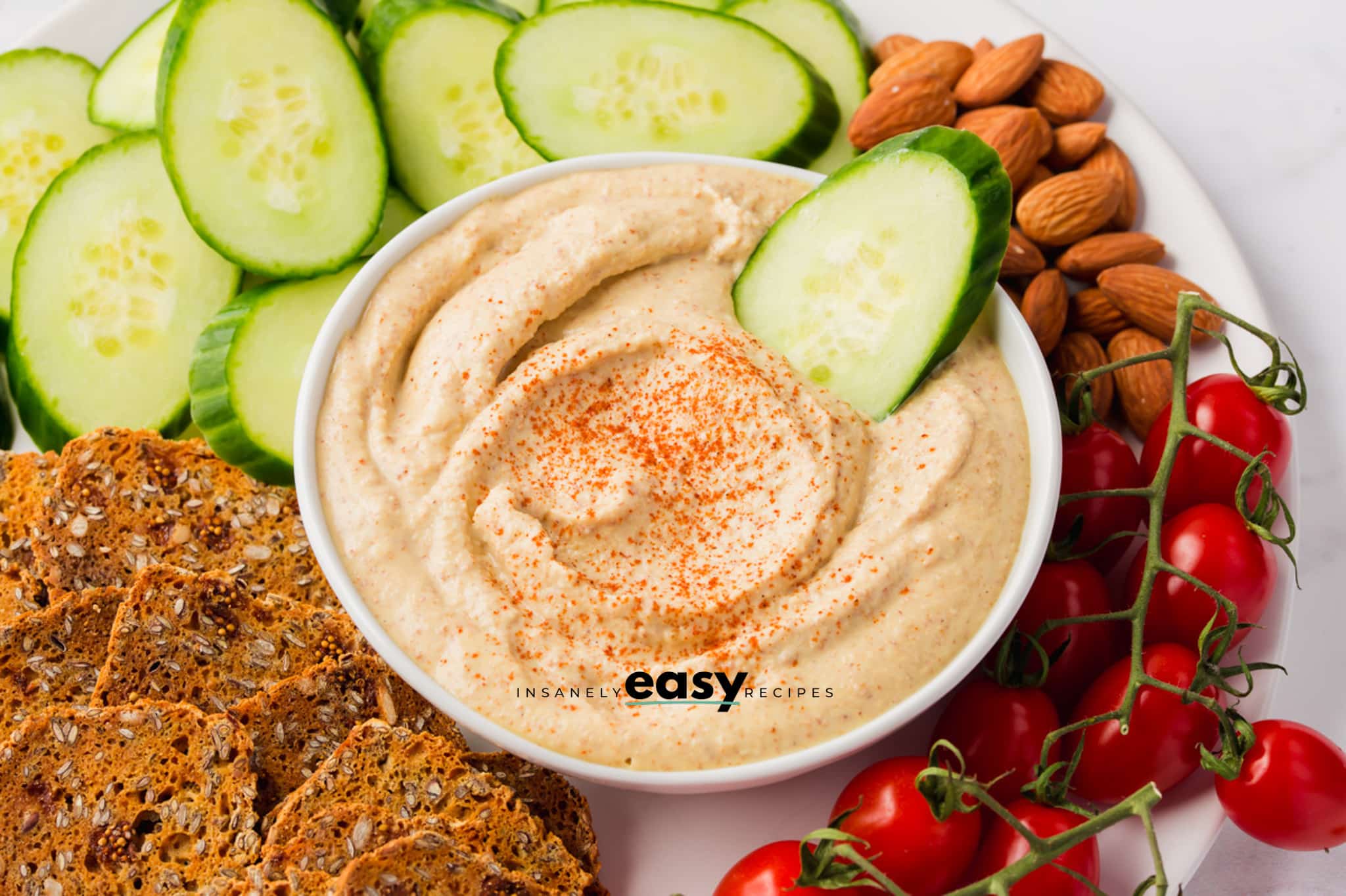 a white bowl of homemade copycat bitchin sauce, swirled with paprika sprinkled on top. almonds, tomatoes, cucumbers, and seed crackers are around the bowl, and there is a cucumber slice in the bowl of dip