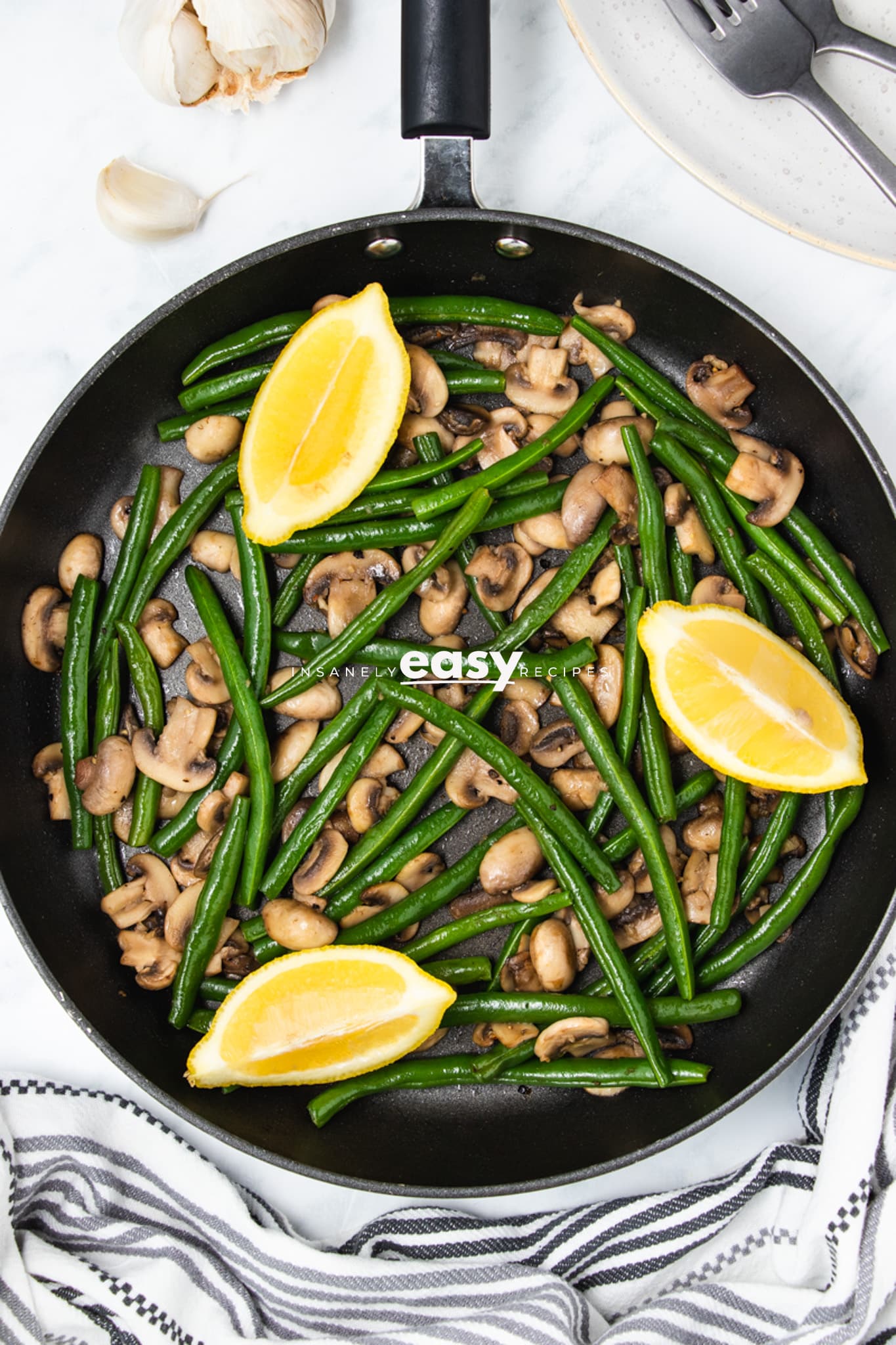 Sauteed green beans and mushrooms in a skillet with lemon wedges on top. The skillet is on a table with fresh garlic, a striped kitchen towel, and a small cutting board around it.