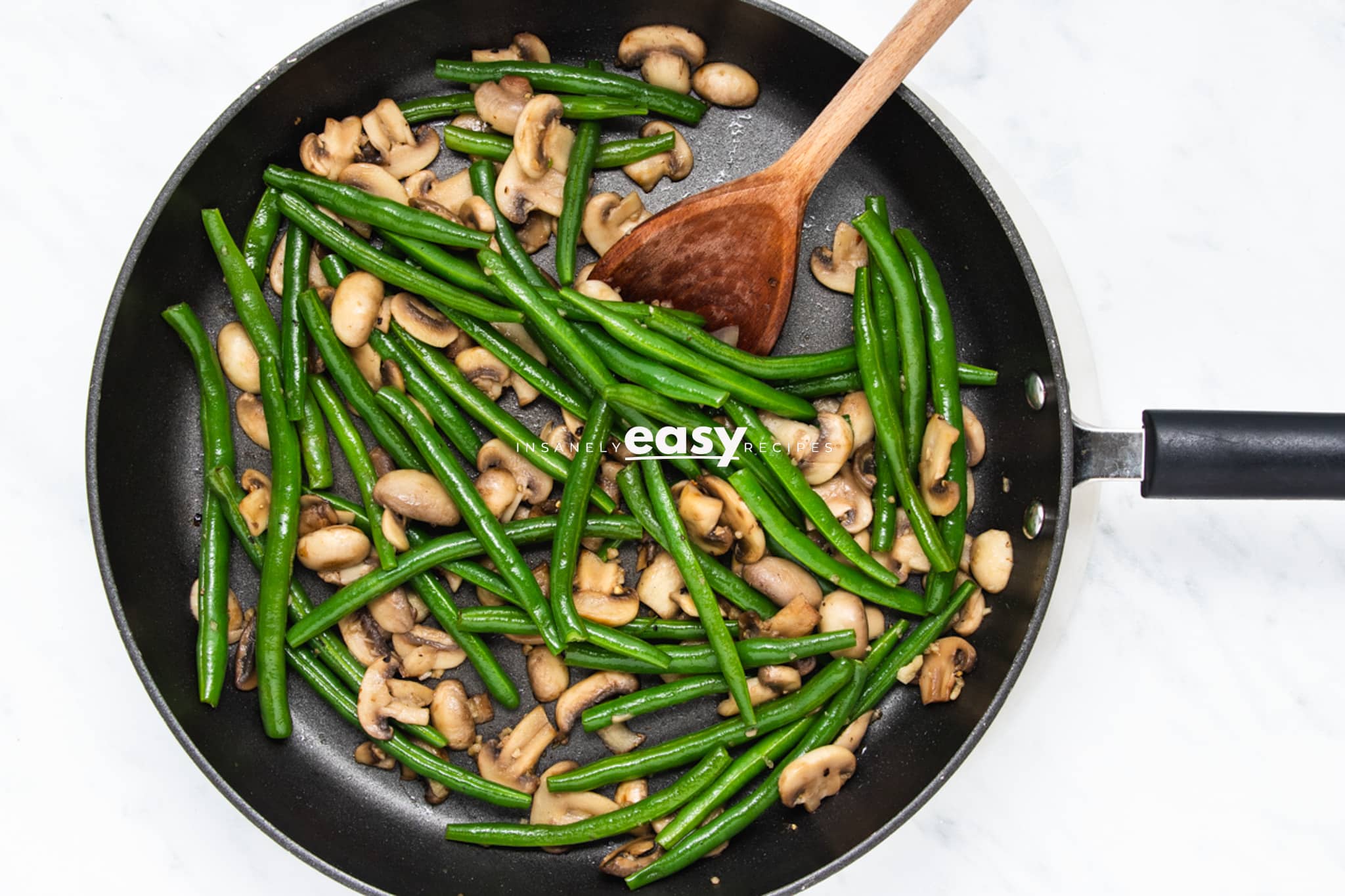 green beans and mushrooms sauteed in a frying pan with a wooden spoon.