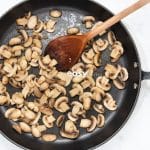 sliced mushrooms sauteed in a frying pan with a wooden spoon.