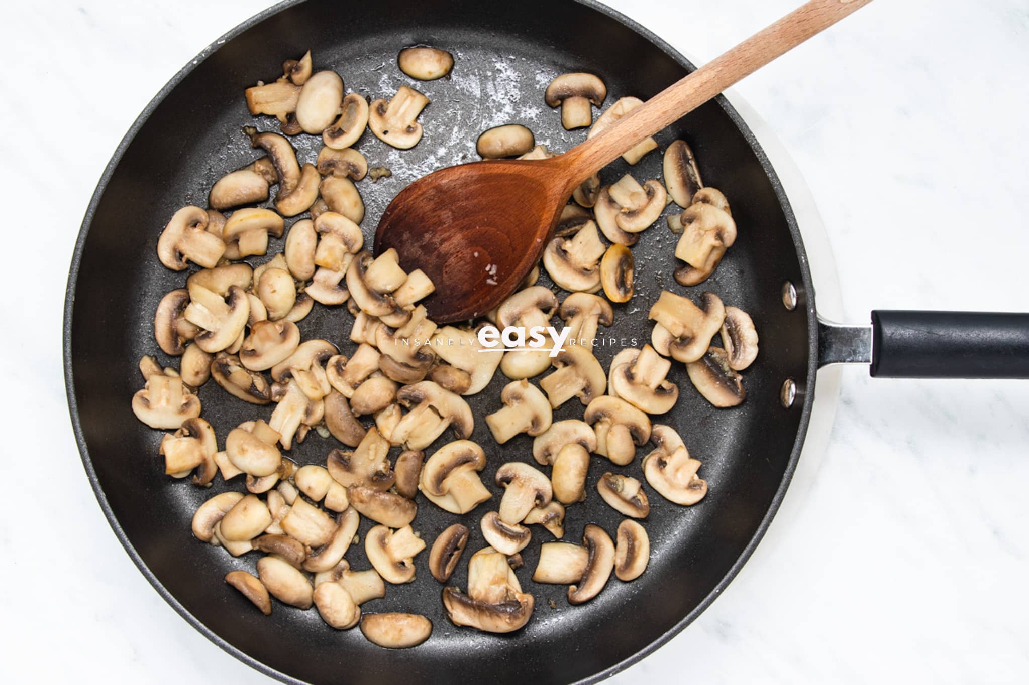 sliced mushrooms sauteed in a frying pan with a wooden spoon.