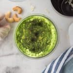 top view photo of cashew pesto that has been blended together. The jar is surrounded by cashews, garlic cloves, and a kitchen towel