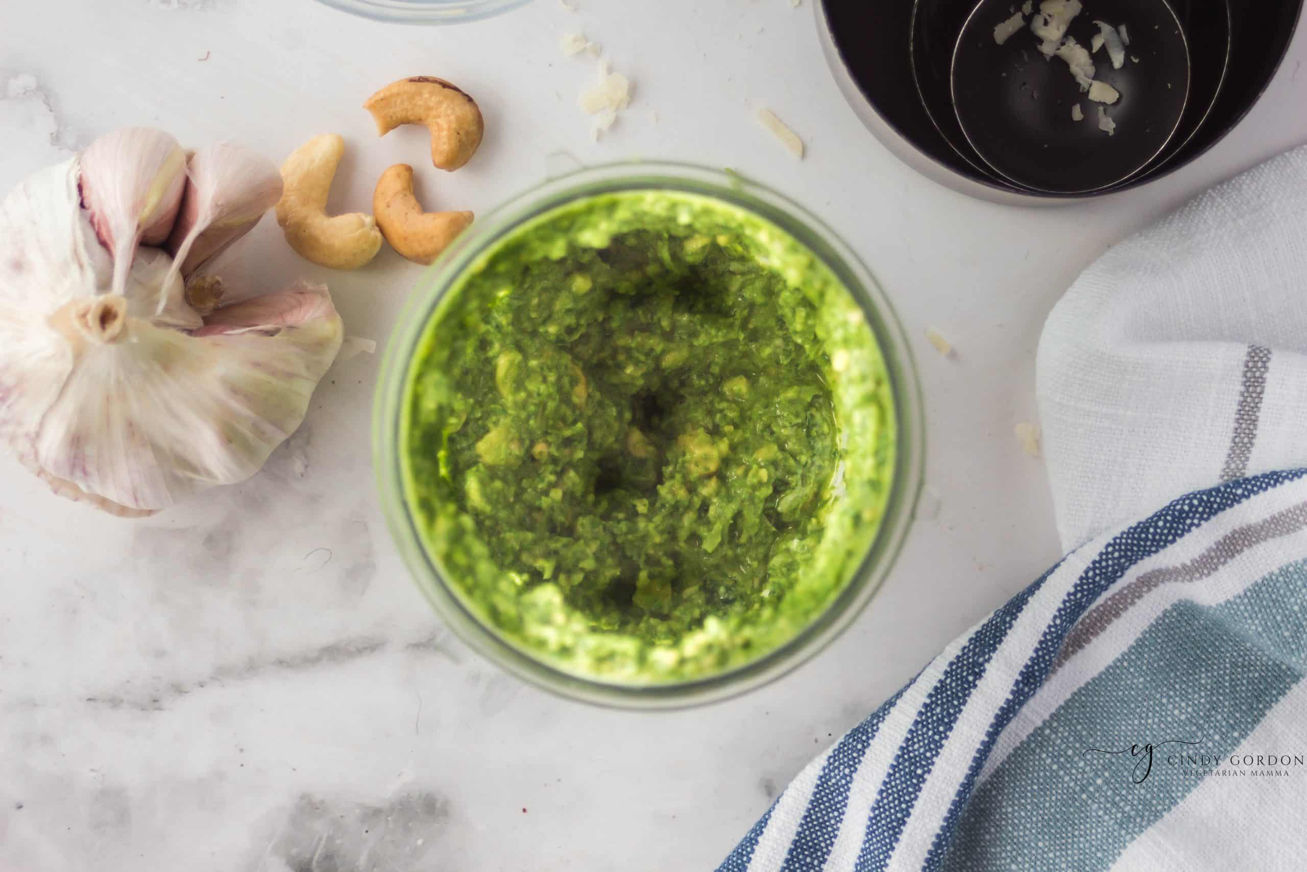 top view photo of cashew pesto that has been blended together. The jar is surrounded by cashews, garlic cloves, and a kitchen towel