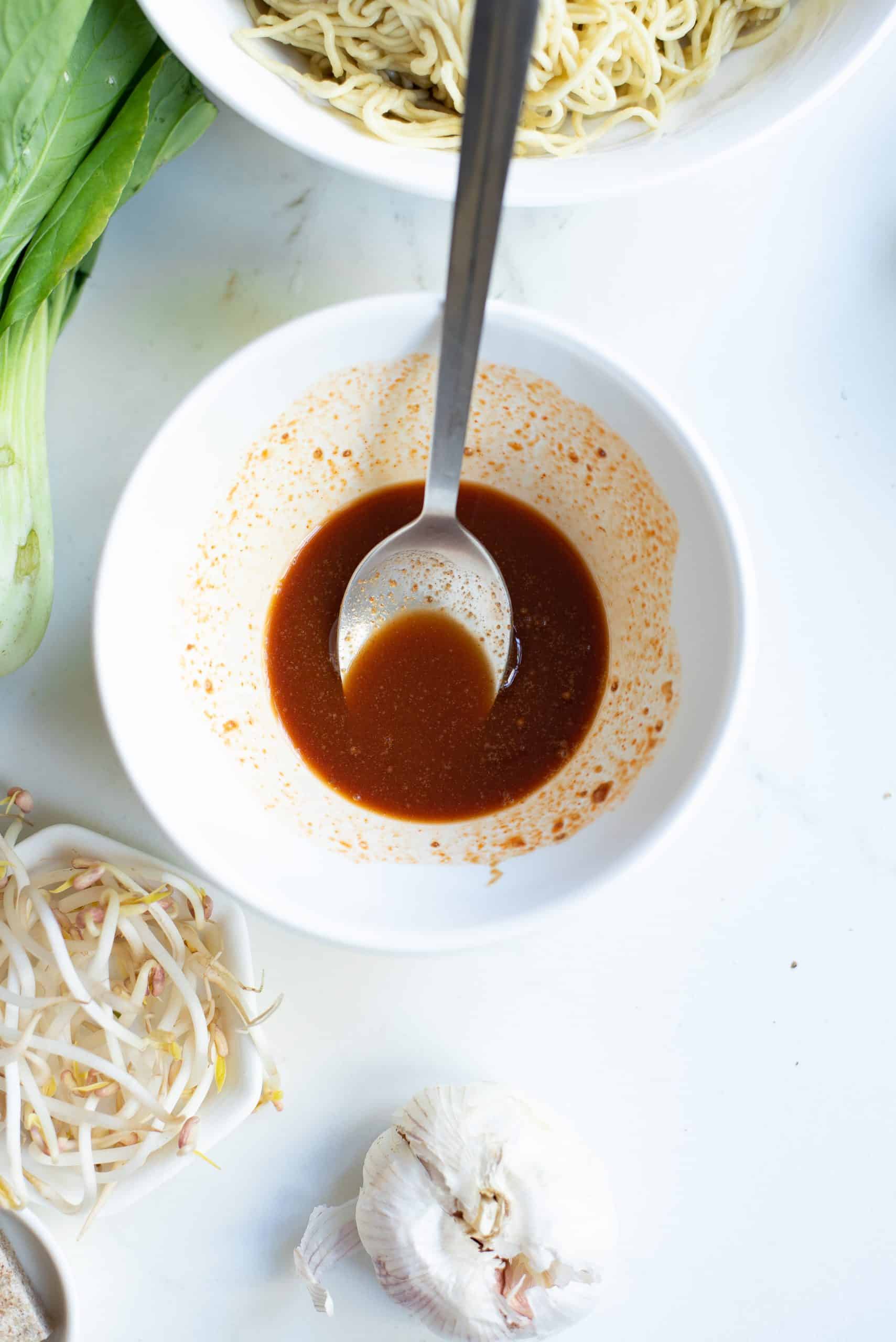 Top view photo of ginger powder, garlic powder, soy sauce, and sriracha mixed together in a white bowl. The bowl is surrounded by garlic, bean sprouts, boy choy, and noodles in a decorative manner.