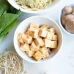 Top view photo of cubed tofu in a white bowl with boy choy, garlic, bean sprouts, mushrooms and noodles around the bowl in a decorative manner.