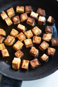 Closeup photo of cubed tofu searing in a cast iron skillet with oil until lightly browned.