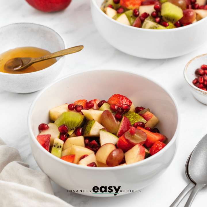 Photo of 2 servings of Thanksgiving Fruit Salad in two white bowls. There is a white bowl with honey and a small white bowl with pomegranate seeds by the salad.