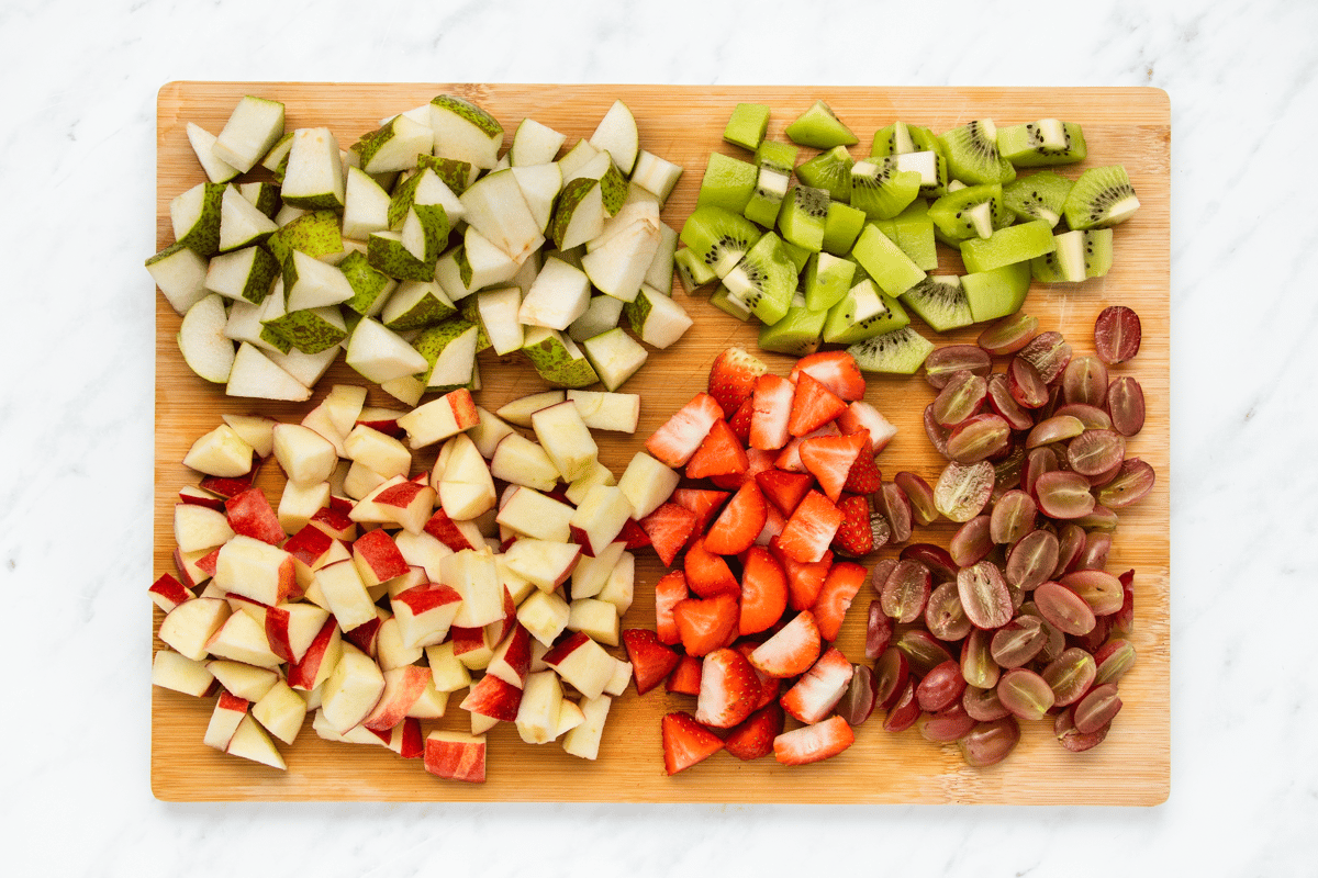 Top view photo of all the fruit that needs to be chopped in Thanksgiving Fruit Salad, on a cutting board, and chopped into bite-sized pieces.