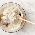 top view photo of gluten free flour, cinnamon, ginger, baking soda, and xanthan gum in a glass bowl with a wooden spoon, ready to be mixed.