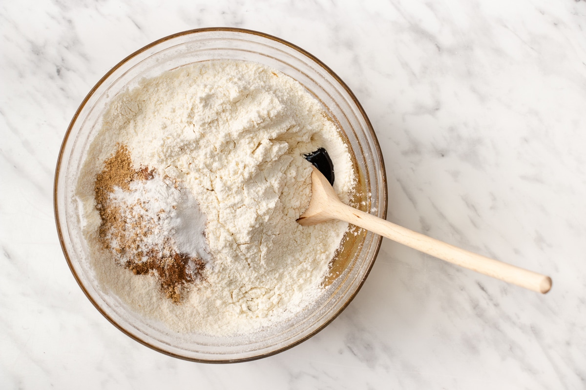 top view photo of gluten free flour, cinnamon, ginger, baking soda, and xanthan gum in a glass bowl with a wooden spoon, ready to be mixed.