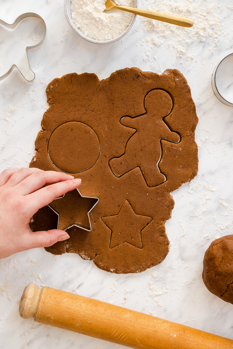 top view photo of gingerbread dough rolled out with a hand using a star cookie cutter to make shapes in the dough. There is a rolling pin below the dough and a bowl of flour with a gold spoon scooping it out and cookie cutter above the dough