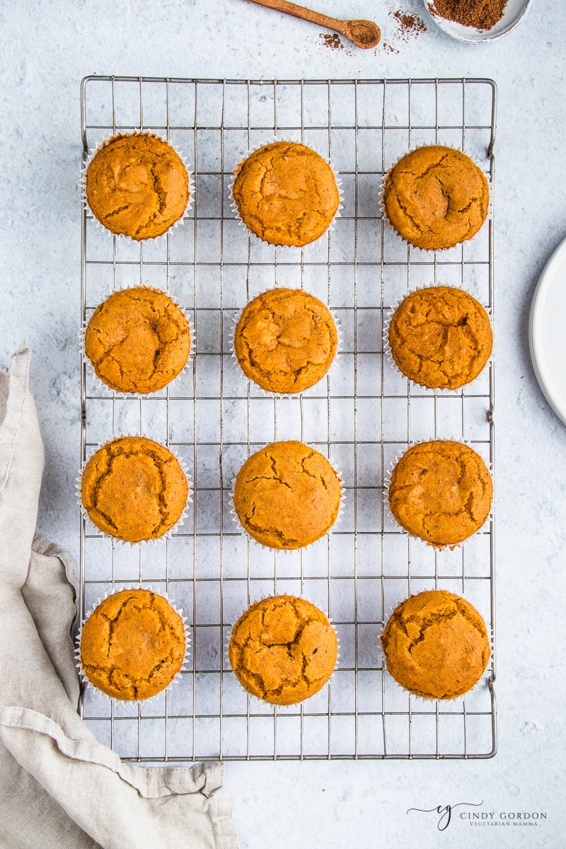 Topview photo of 12 Vegan Pumpkin Muffins, cooling on a wire rack. There is a kitchen towel to the left of the rack and pumpkin spice above the rack. 