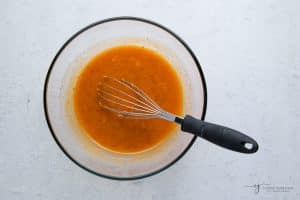 Topview photo of more wet ingredients to make Vegan Pumpkin Muffins, in a clear bowl, being mixed by a wire whisk.