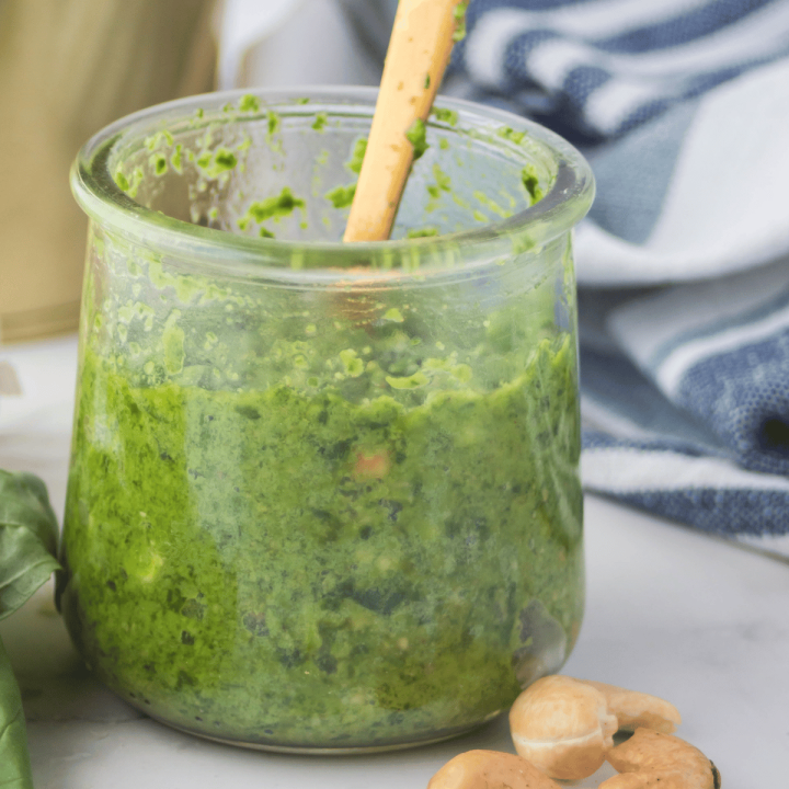 closeup photo of cashew pesto in a small glass jar, with a gold spoon dipped inside. There is a kitchen towel in the background and basil leaves and cashews in the foreground