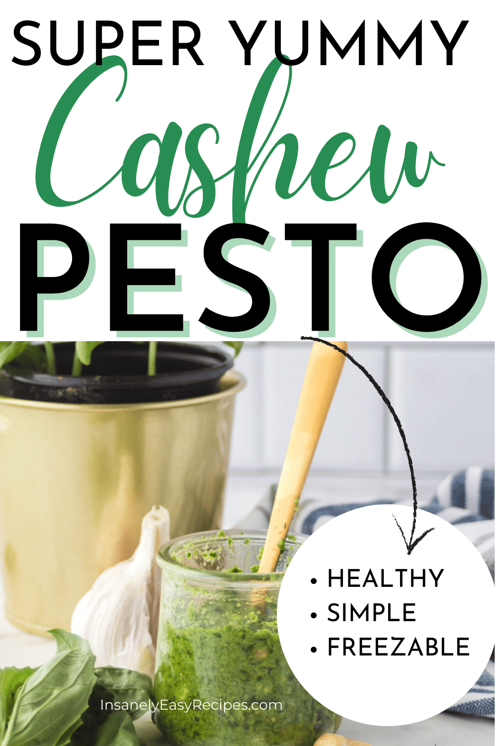 pinterest photo of cashew pesto, in a small glass jar with a gold spoon dipped inside. There is a gold pot of basil and a kitchen towel in the background, and basil, cashews, and garlic in the foreground
