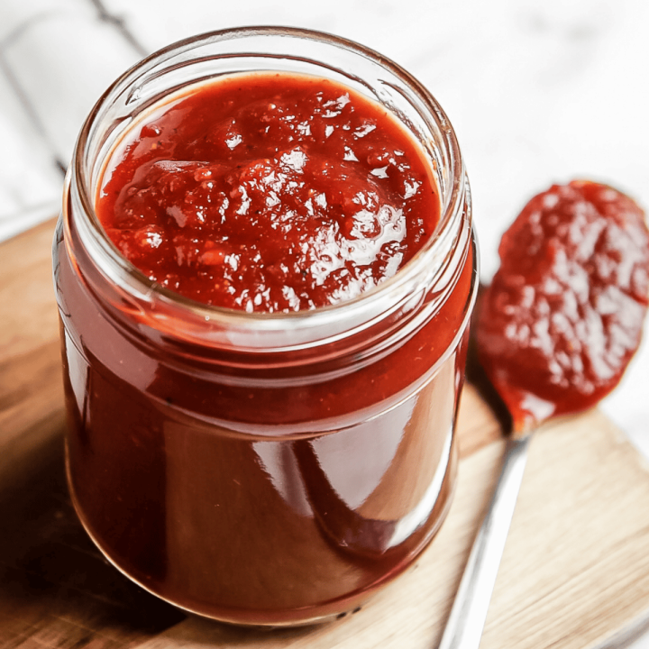 Closeup photo of Vegan BBQ Sauce in a clear jar with a spoon filled with sauce next to the jar.