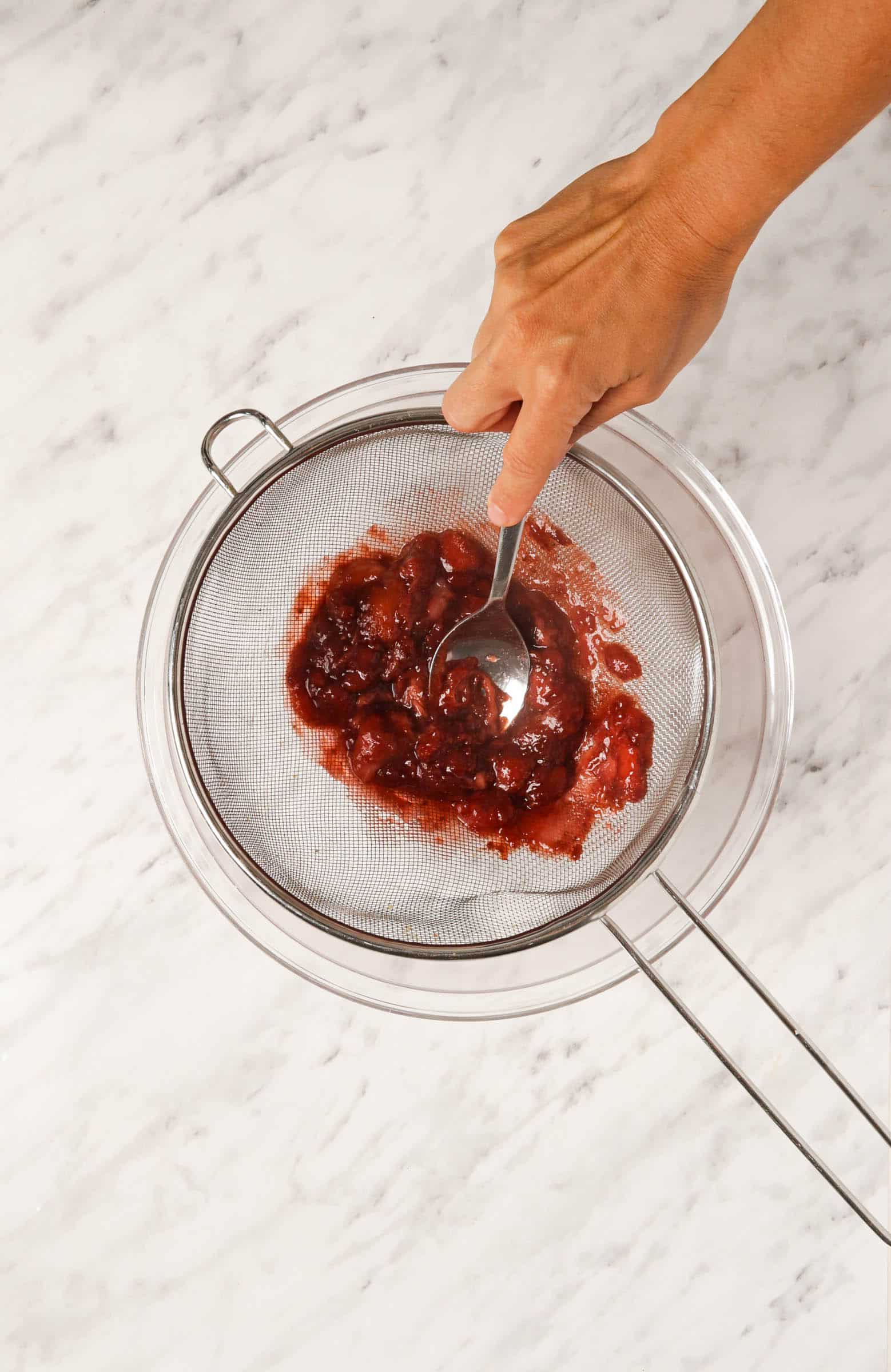 Top view photo of the strawberry mixture in a fine mesh strainer over a bowl. A hand is pressing down on the mixture with the back of a spoon.