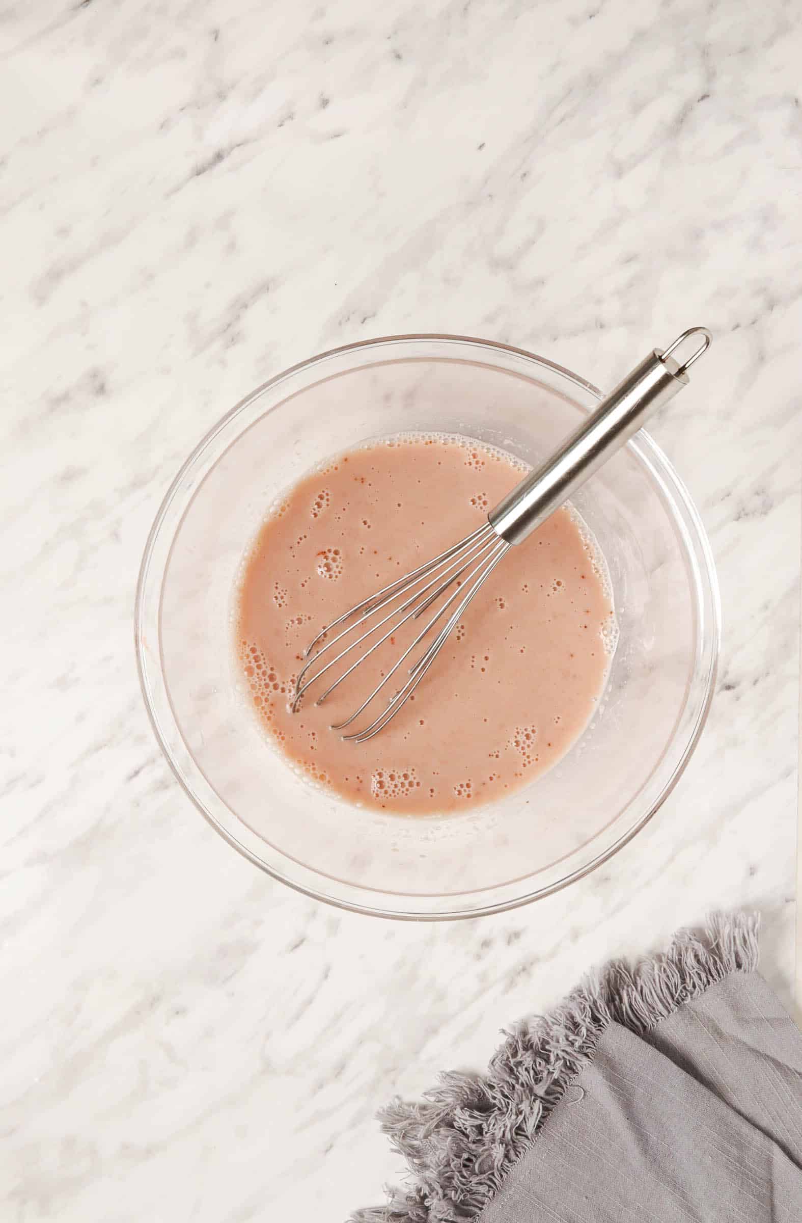 Top view photo of almond milk mixed together with the strawberry mixer with a whisk, in a clear bowl.