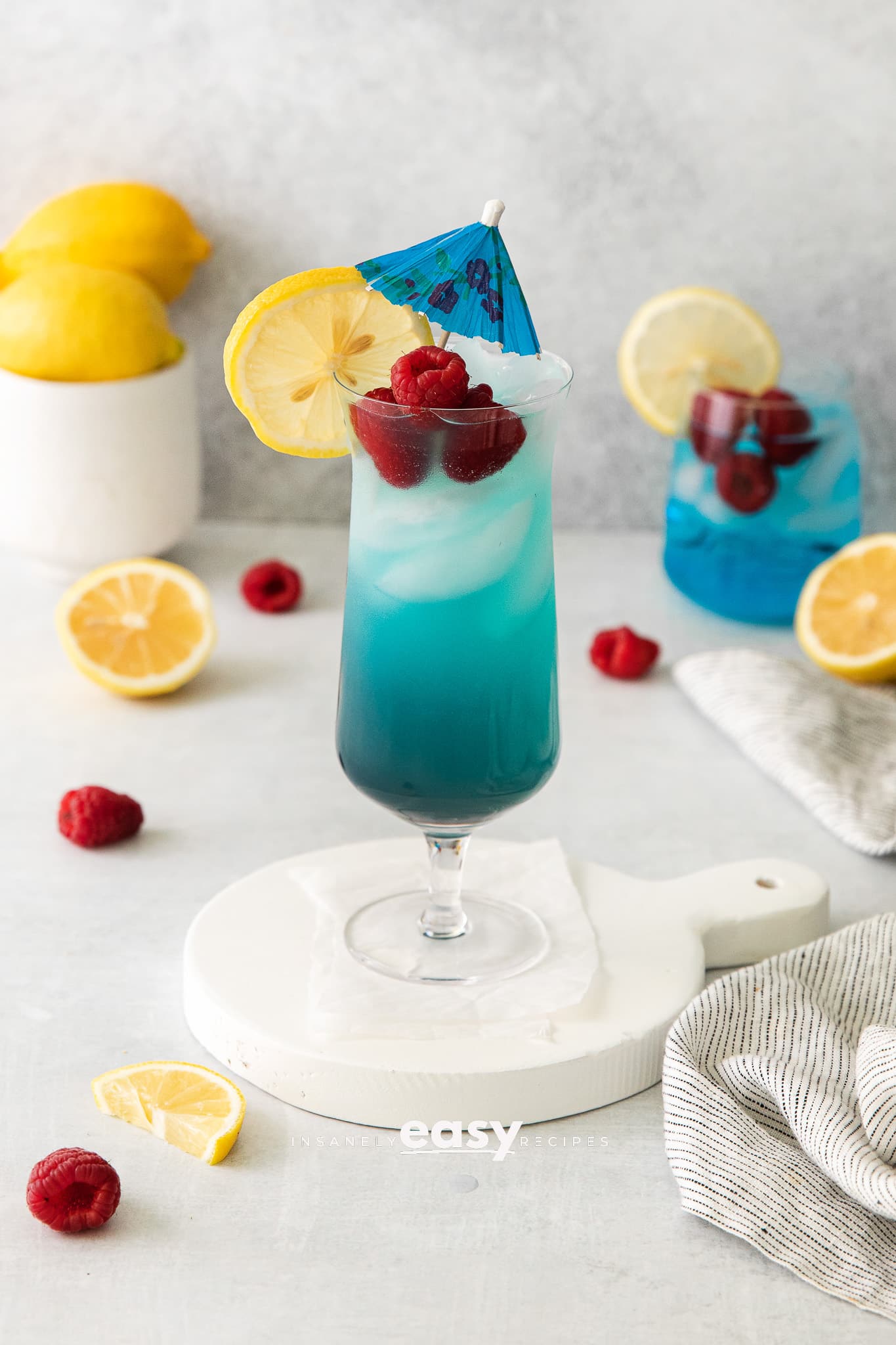 tall clear glass with red liquid at bottom, then blue liquid, with rapsberries and blue umbrella in drink. Surrounded by lemons and raspberries on the table