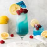 tall clear glass with red liquid at bottom, then blue liquid, with rapsberries and blue umbrella in drink. Surrounded by lemons and raspberries on the table
